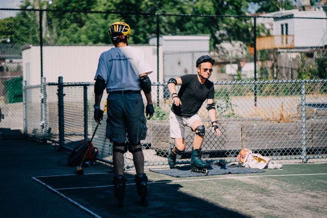 Person in inline skates and protective gear sitting on pavement next to chain-link fence