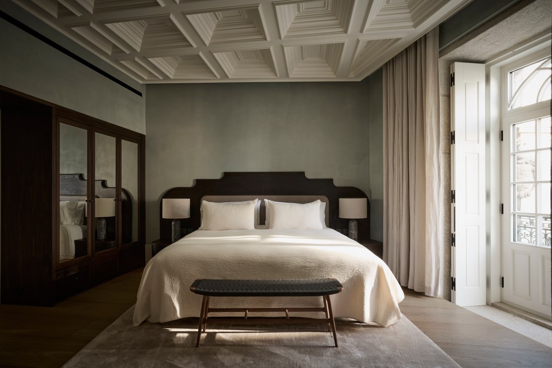 Elegant hotel bedroom with coffered ceiling, dark wood headboard, white linens, and French doors opening to balcony