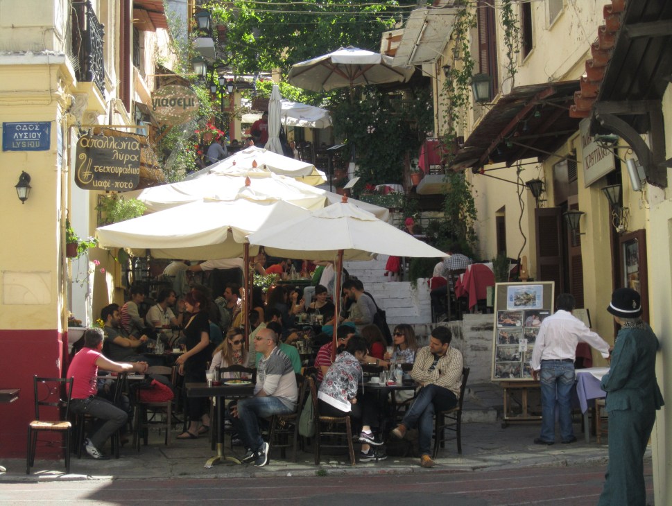 People dining at outdoor cafe tables with white umbrellas on a narrow street in Athens