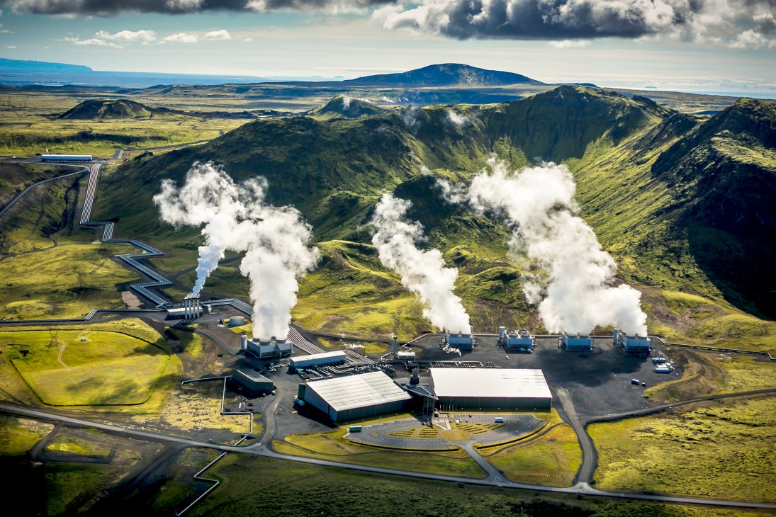 Aerial view of Hellisheidi geothermal power plant in Iceland with steam plumes rising from facilities in volcanic landscape.