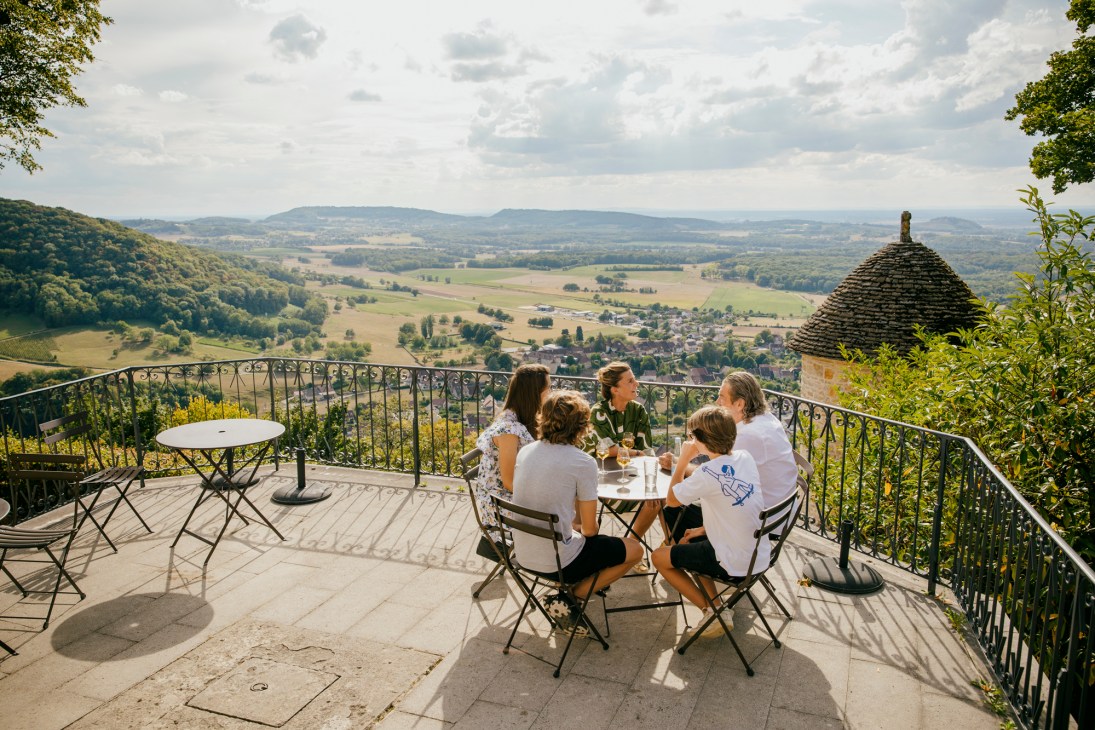 Group of people dining on terrace overlooking valley landscape with stone building and mountains in background