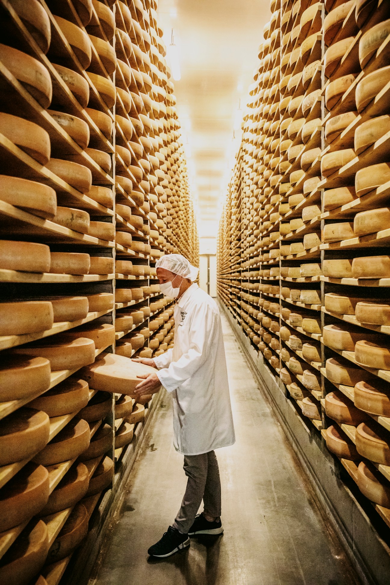 Worker in white coat inspecting wheels of aging comté cheese on wooden shelves in Fromageries Arnaud cellar