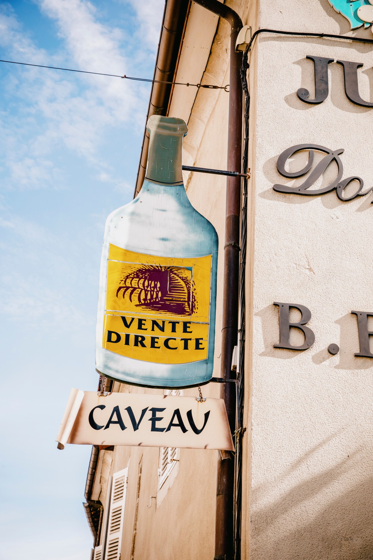 Wine bottle sign with "VENTE DIRECTE" and "CAVEAU" text mounted on building exterior in Poligny