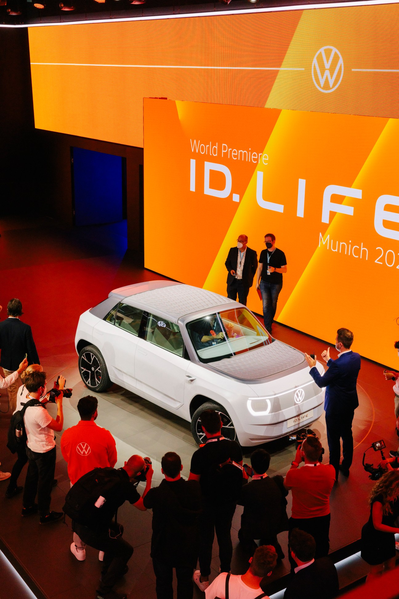 Volkswagen ID.Life concept car on display at IAA Munich with crowds and orange branding backdrop.