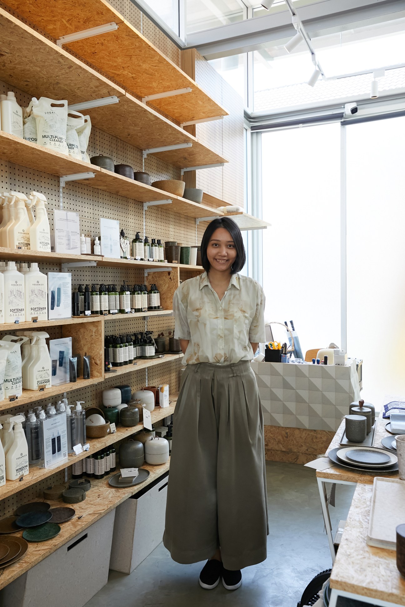 Woman in cream shirt standing in modern showroom with wooden shelving displaying home goods and cleaning products.