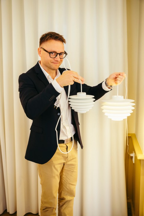 Man in black blazer and glasses holding white pendant lamps with cord details against cream curtains.