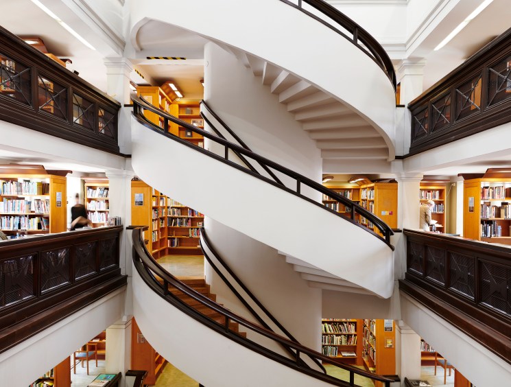 White spiral staircase with dark railings curves through multi-level library with bookshelves and warm lighting
