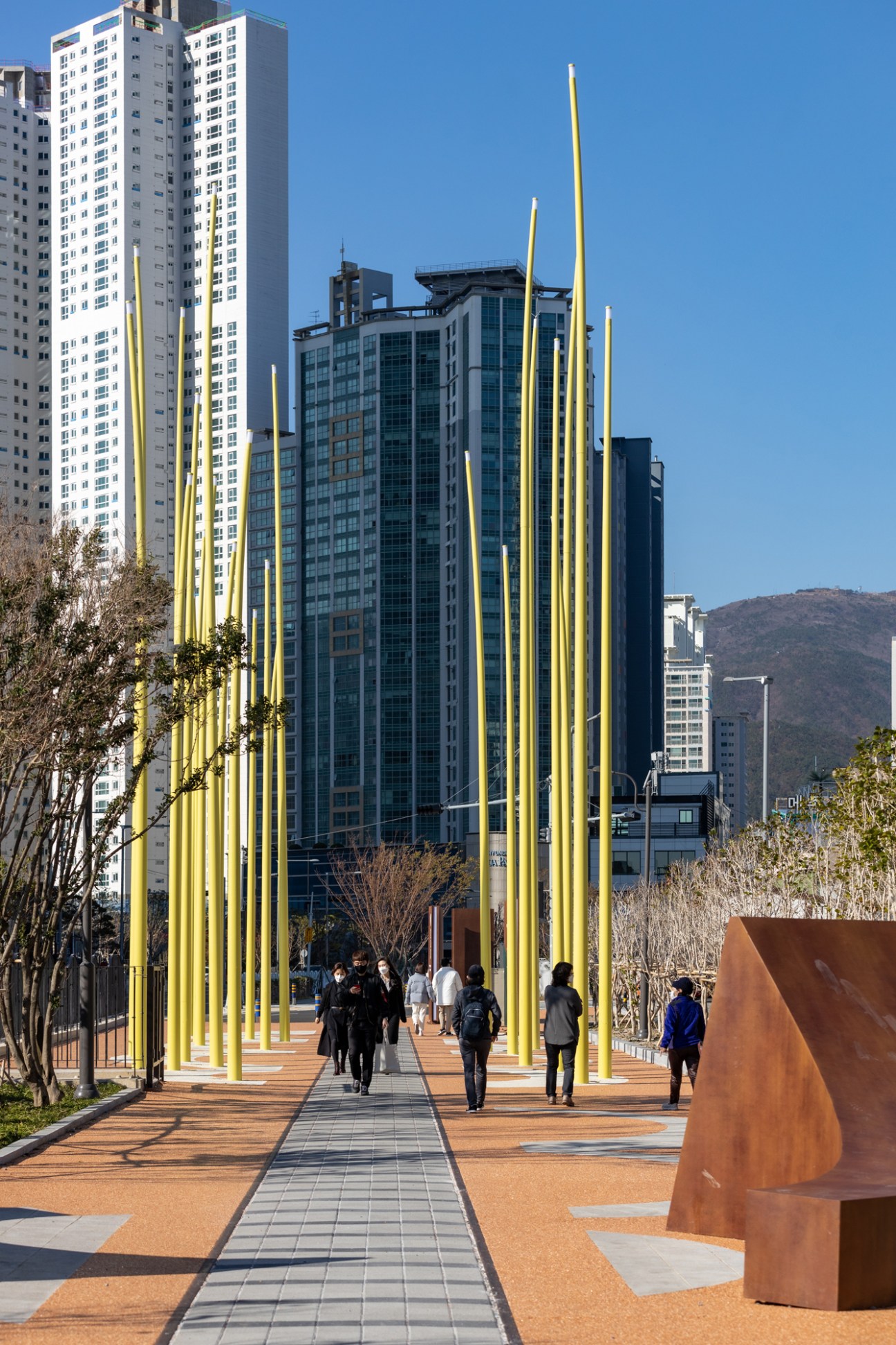 Modern urban park walkway with tall yellow poles and people strolling, buildings and mountains in background