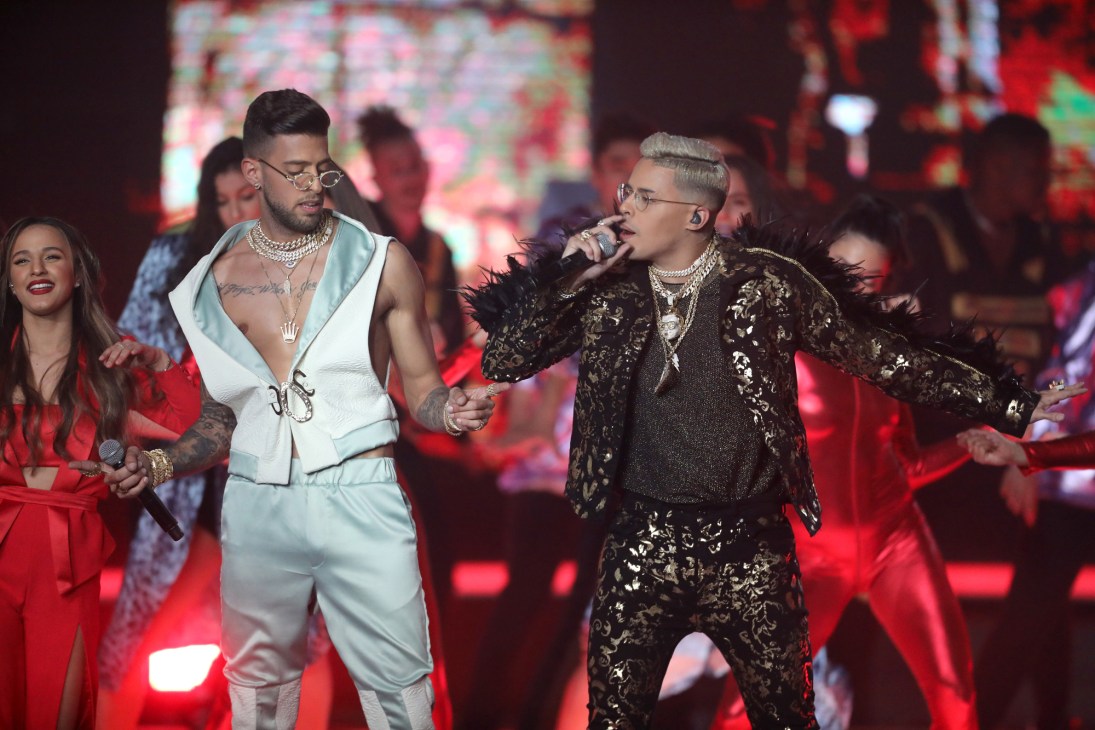 Two male performers singing on stage, one in light blue outfit, one in dark patterned clothing with microphones