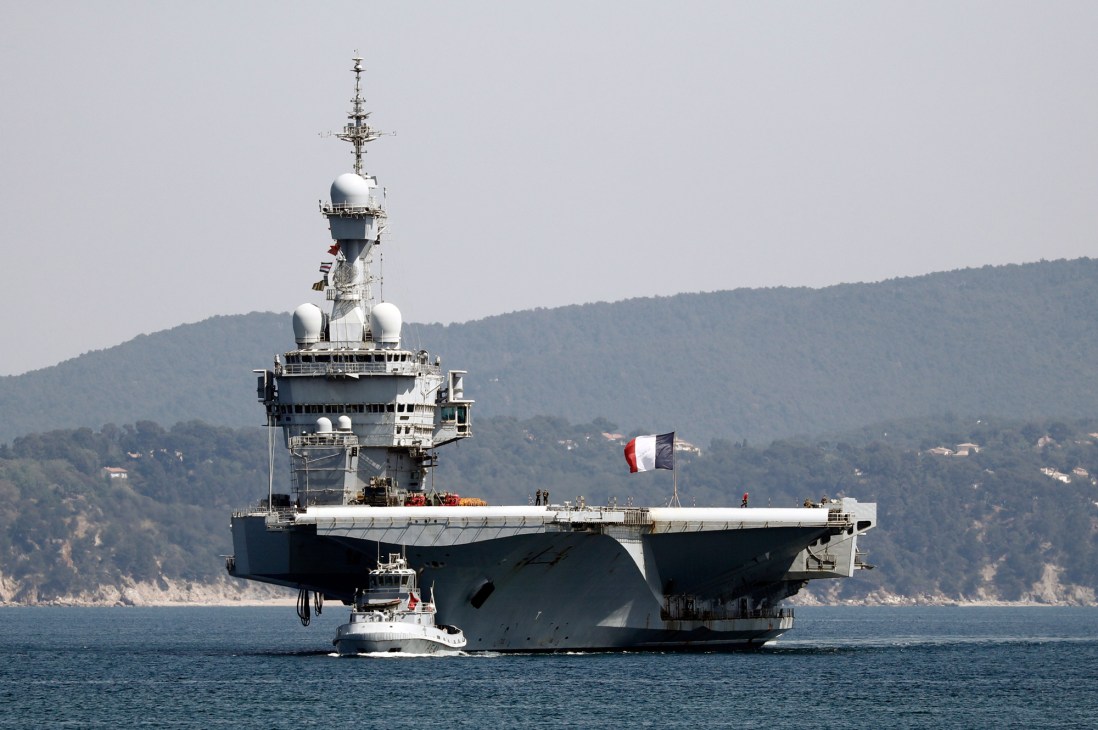 French aircraft carrier Charles de Gaulle at sea with French flag flying and mountainous coastline in background.