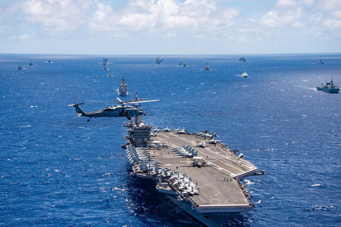 Aircraft carrier with helicopter flying overhead surrounded by naval fleet in blue ocean waters