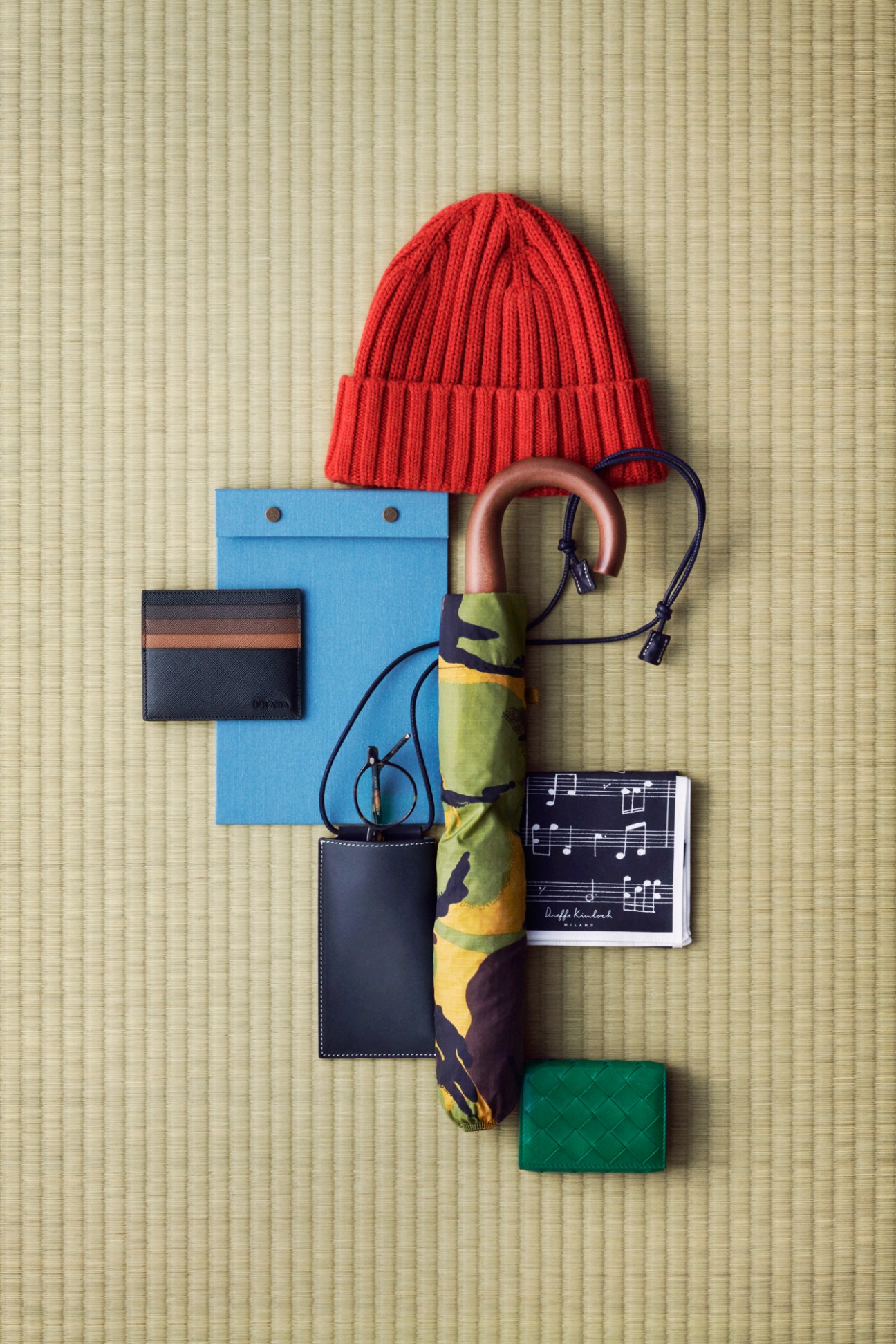 Red knitted beanie, blue notepad, colorful umbrella, patterned handkerchief, card cases and eyeglasses arranged on tatami mat