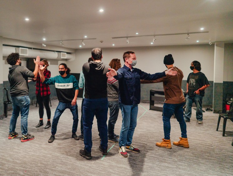 Group of masked people in casual clothing standing in a circle doing improv exercises in a modern classroom