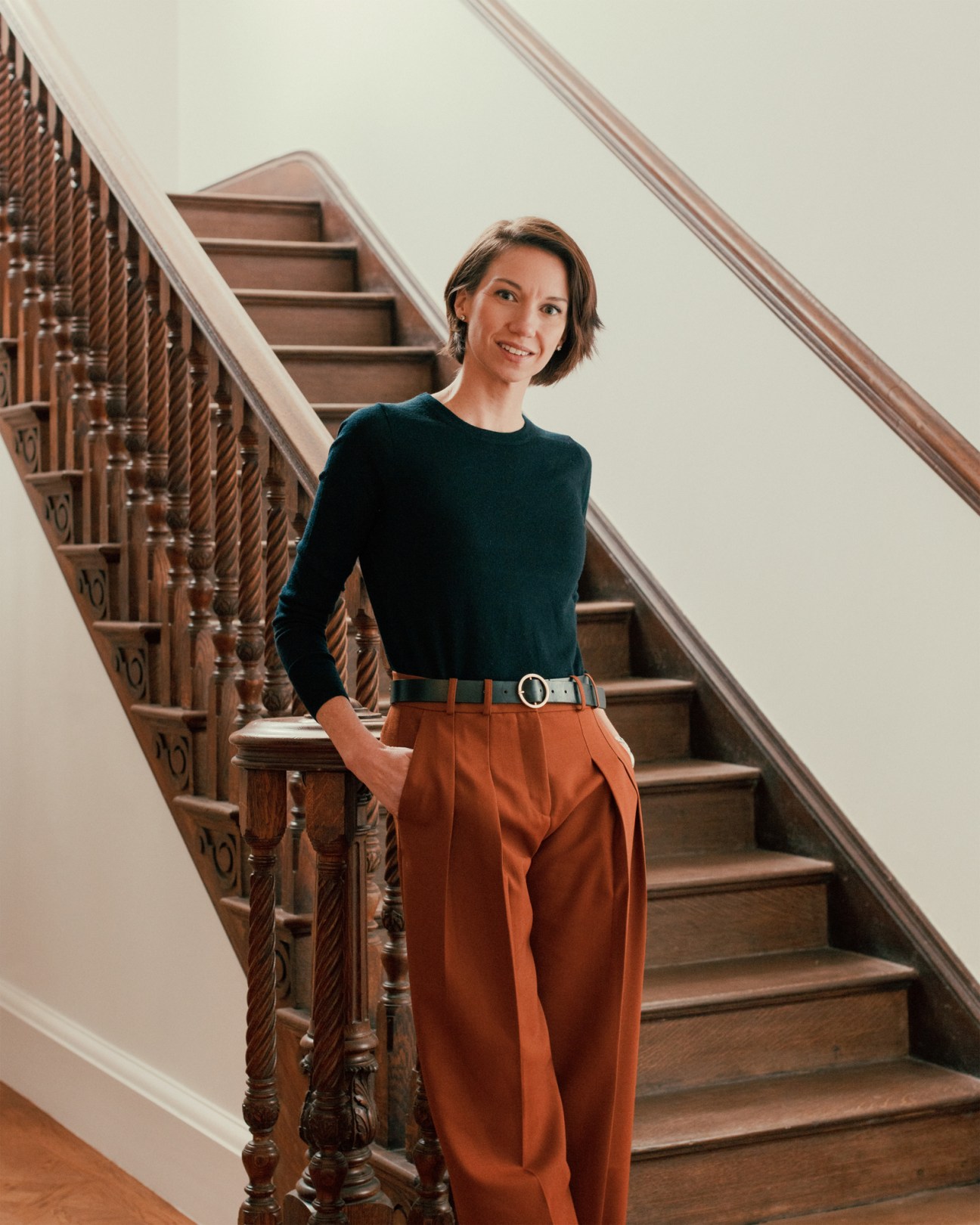 Woman in dark green sweater and brown pants standing by ornate wooden staircase in elegant interior