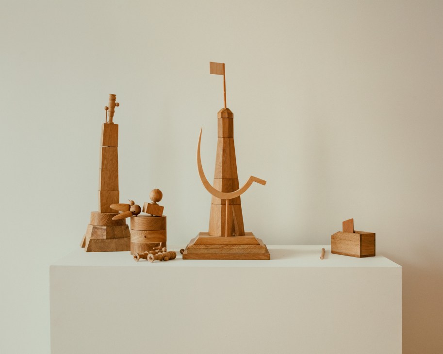 Wooden sculptural artwork with geometric blocks, towers, and curved elements arranged on white surface