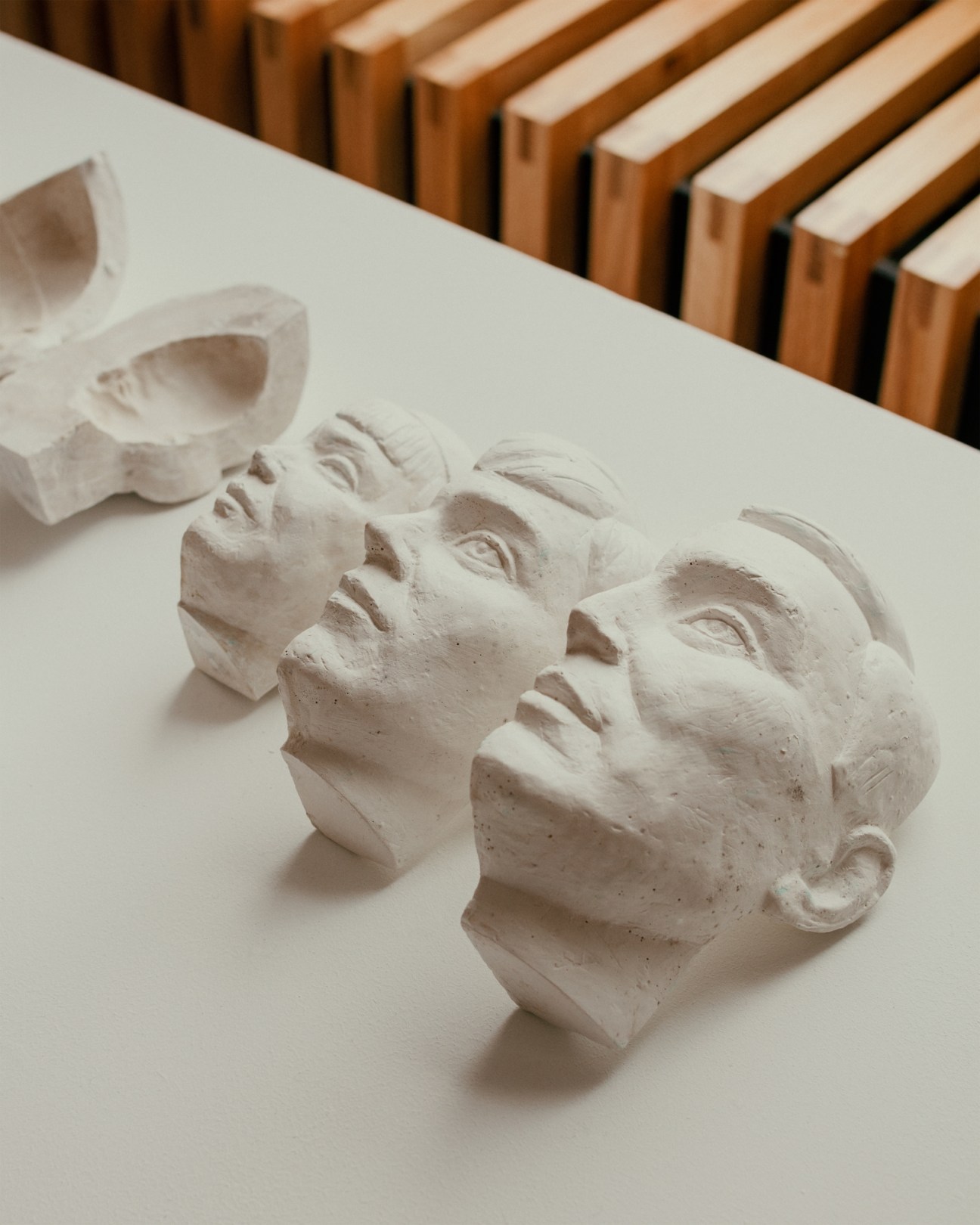 Three white plaster sculptural busts or heads displayed on a white surface with wooden frames in background