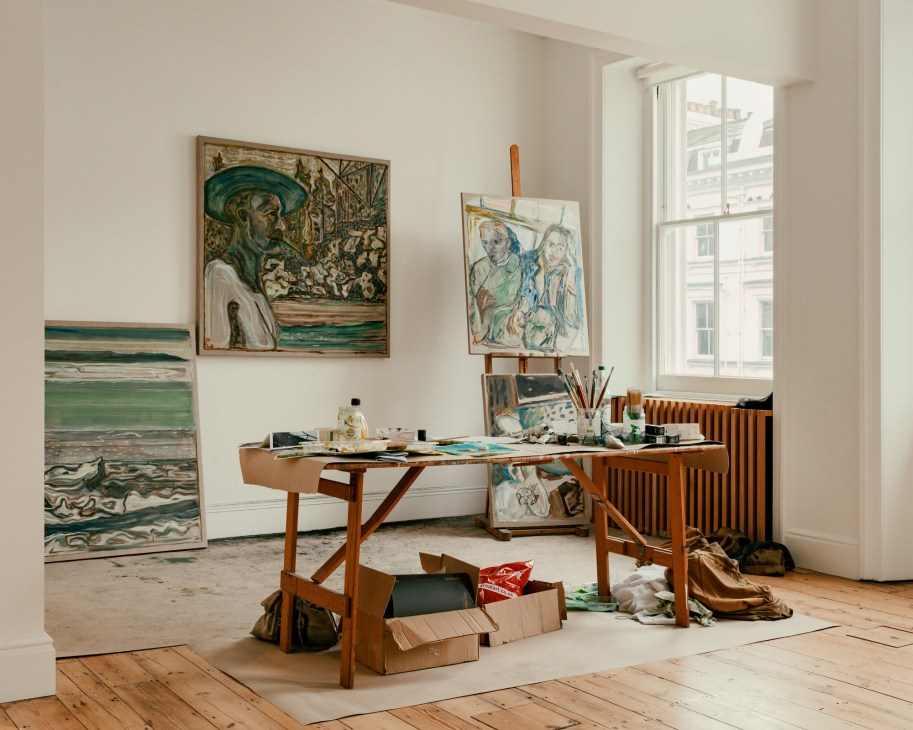 Artist studio with easel, paintings, and art supplies in a bright room with wooden floors and large window