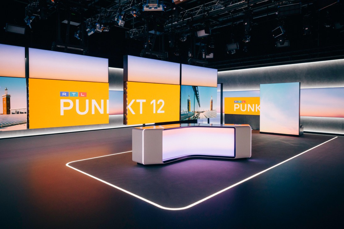 Modern television news studio with curved white anchor desk, large screens displaying orange RTL branding, and lighting rig