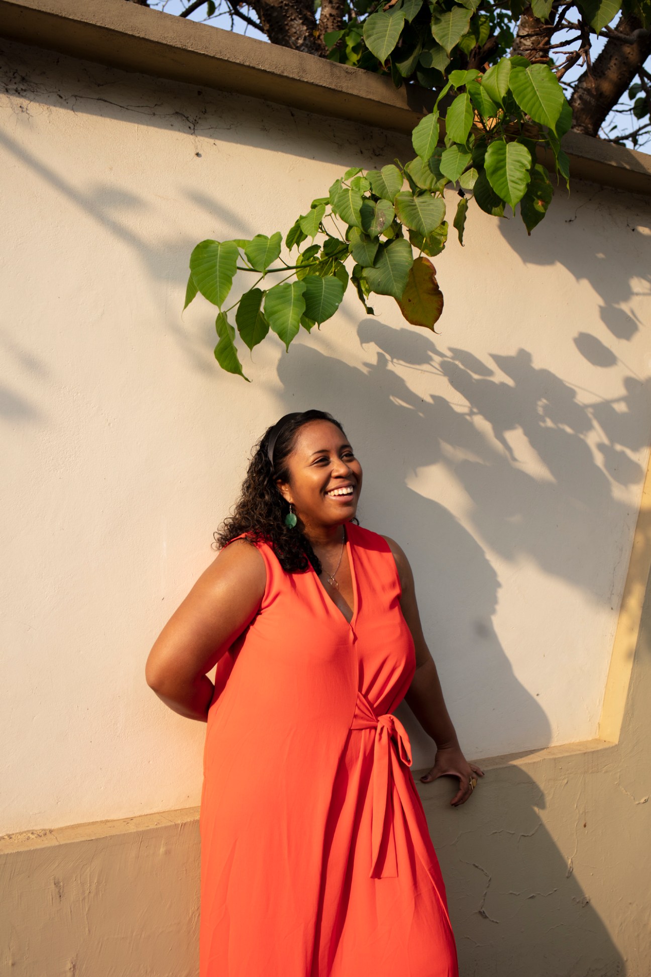 Mae-ling Lokko in an orange wrap dress smiling outdoors against a white wall with leafy shadows