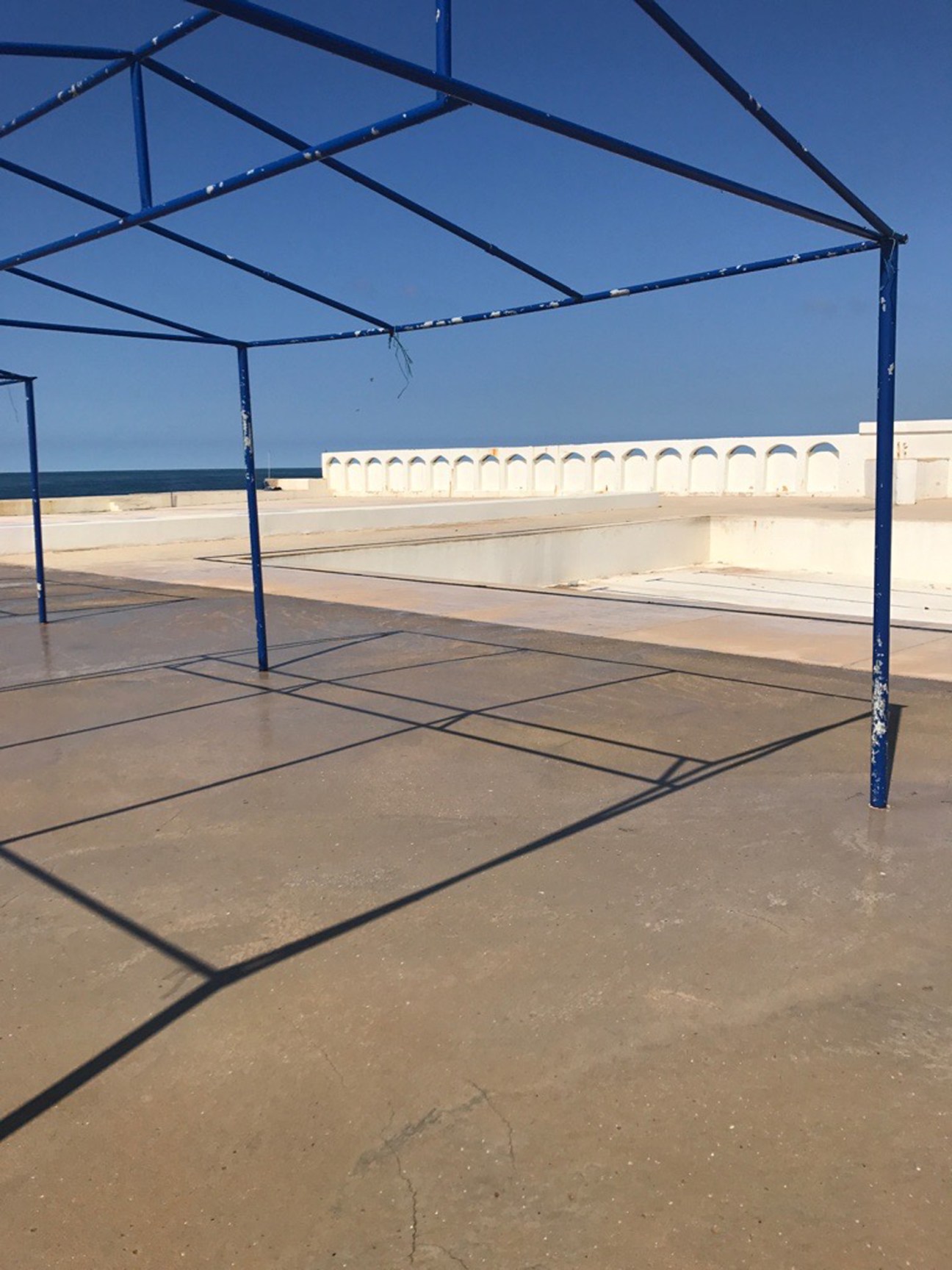 Blue metal frame structure on concrete terrace overlooking sea with white balustrade at Sporting Club Beach