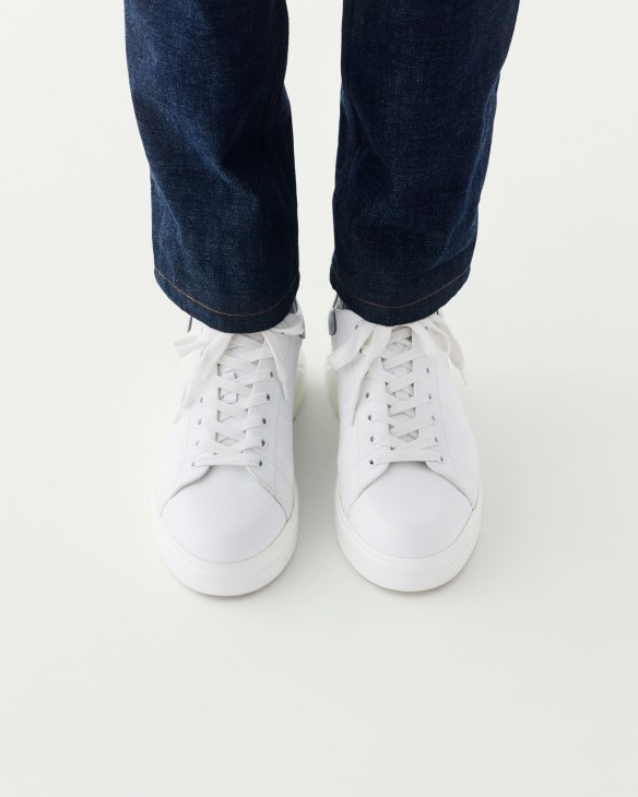 Person wearing dark jeans, gray socks, and white lace-up sneakers standing against a light background.