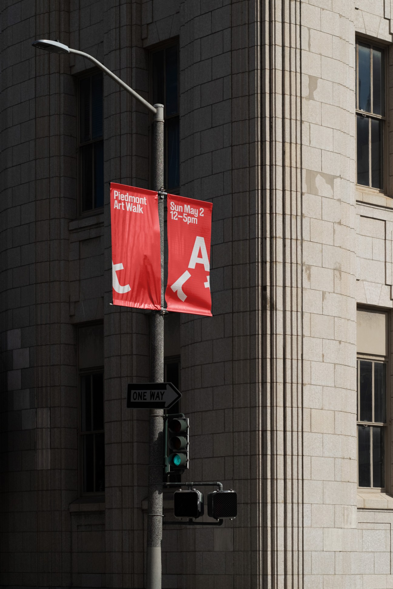 Red banner for Piedmont Art Walk hanging from street lamp pole, featuring stylized letters forming a cute mascot character, with "Sun May 2 12-5pm" and event details on dark urban building backdrop