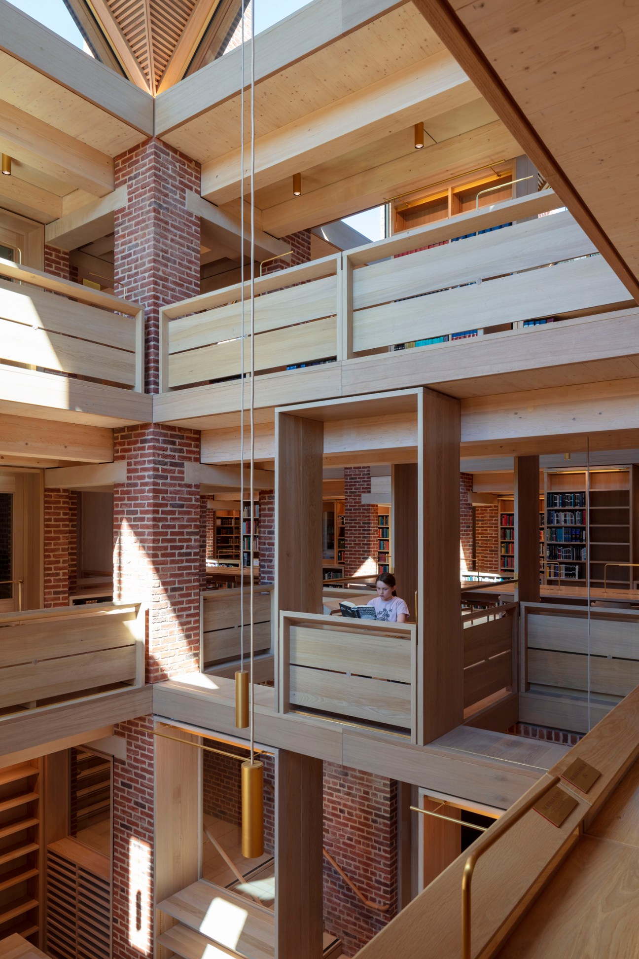 Multi-level library interior with wooden structure, brick columns, and person reading at desk surrounded by bookshelves