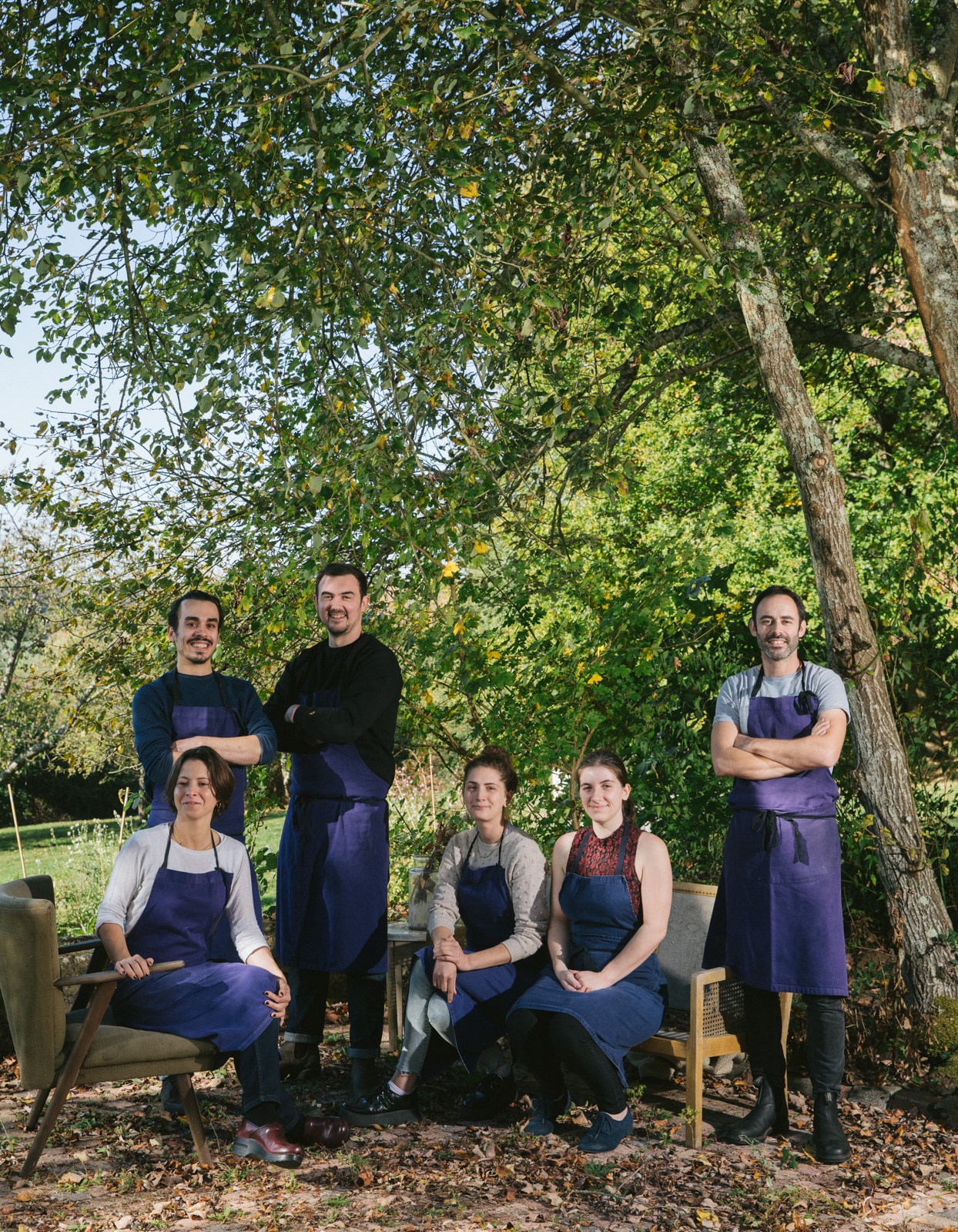 Seven staff members wearing purple aprons pose in a group under large green trees at D'une île in Rémalard, France.