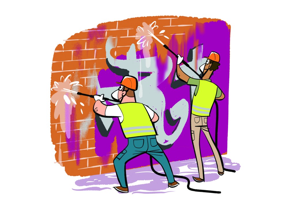 Two workers in high-vis vests and hard hats cleaning graffiti from a brick wall using long-handled tools
