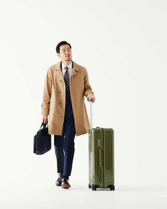 Man in beige coat and navy suit carrying a Rimowa suitcase and Briefing bag.