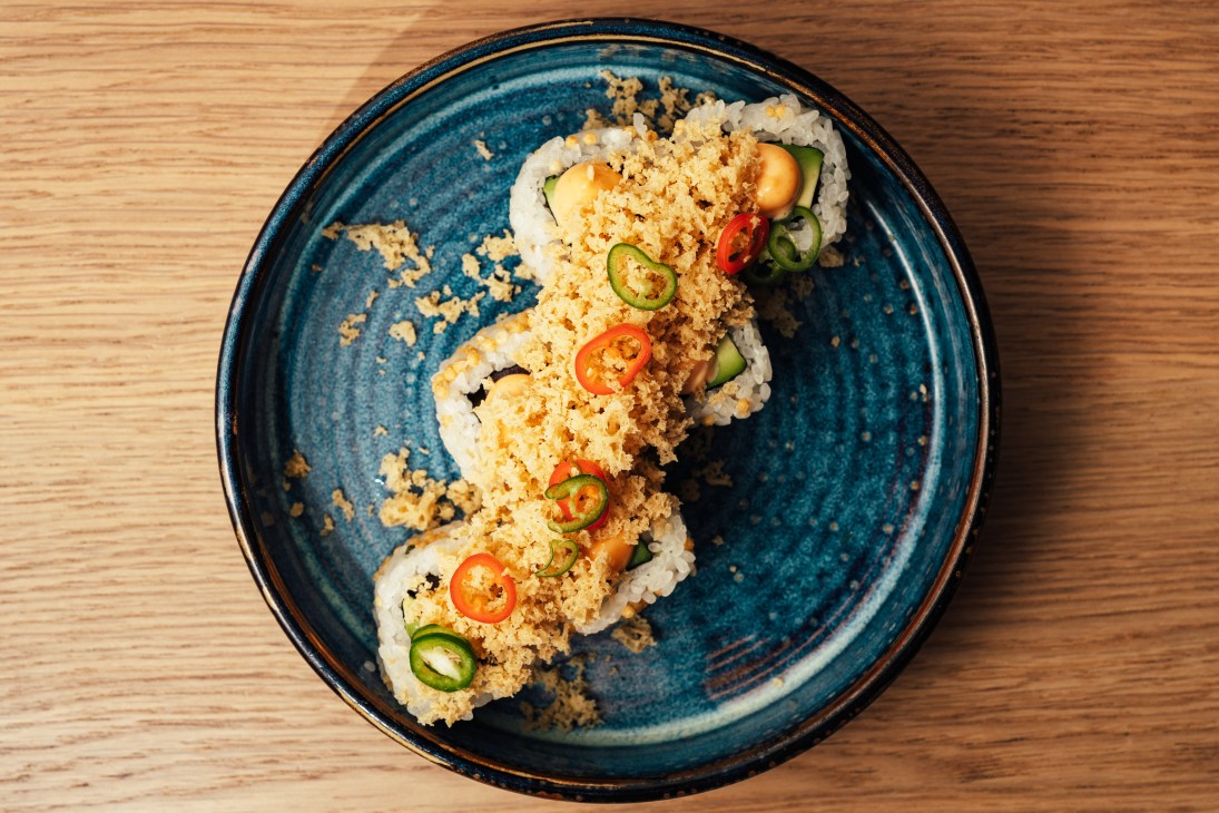 Sushi rolls topped with crispy coating, sliced chili and cucumber on a blue ceramic plate.