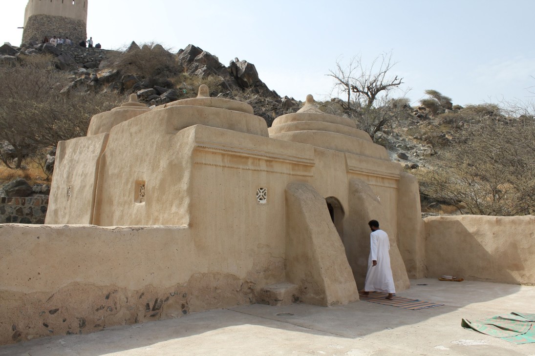 Al Bidya mosque with traditional mud and stone architecture, person in white robe walking near entrance