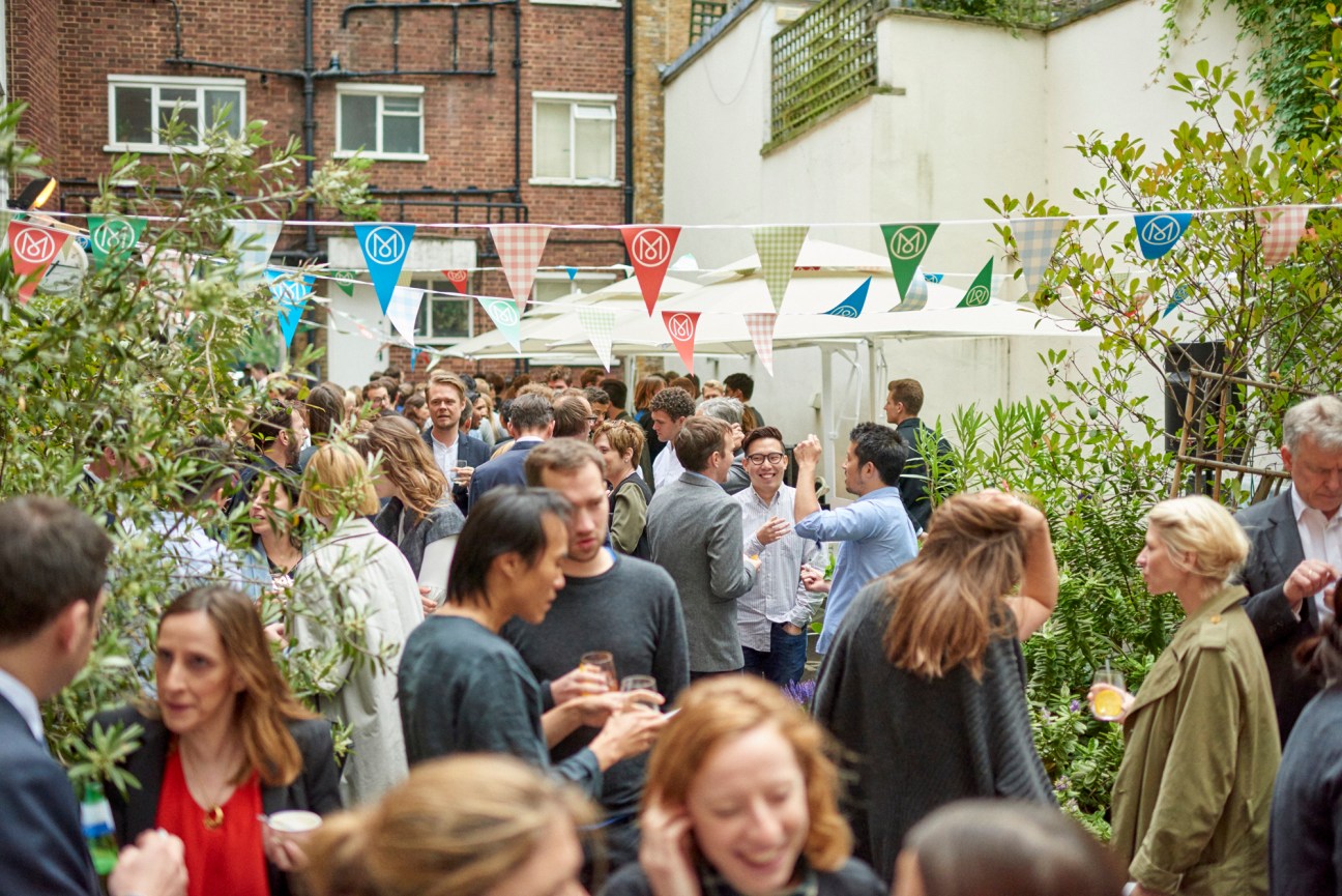 Crowded outdoor party with people mingling under colorful triangular bunting in a garden courtyard