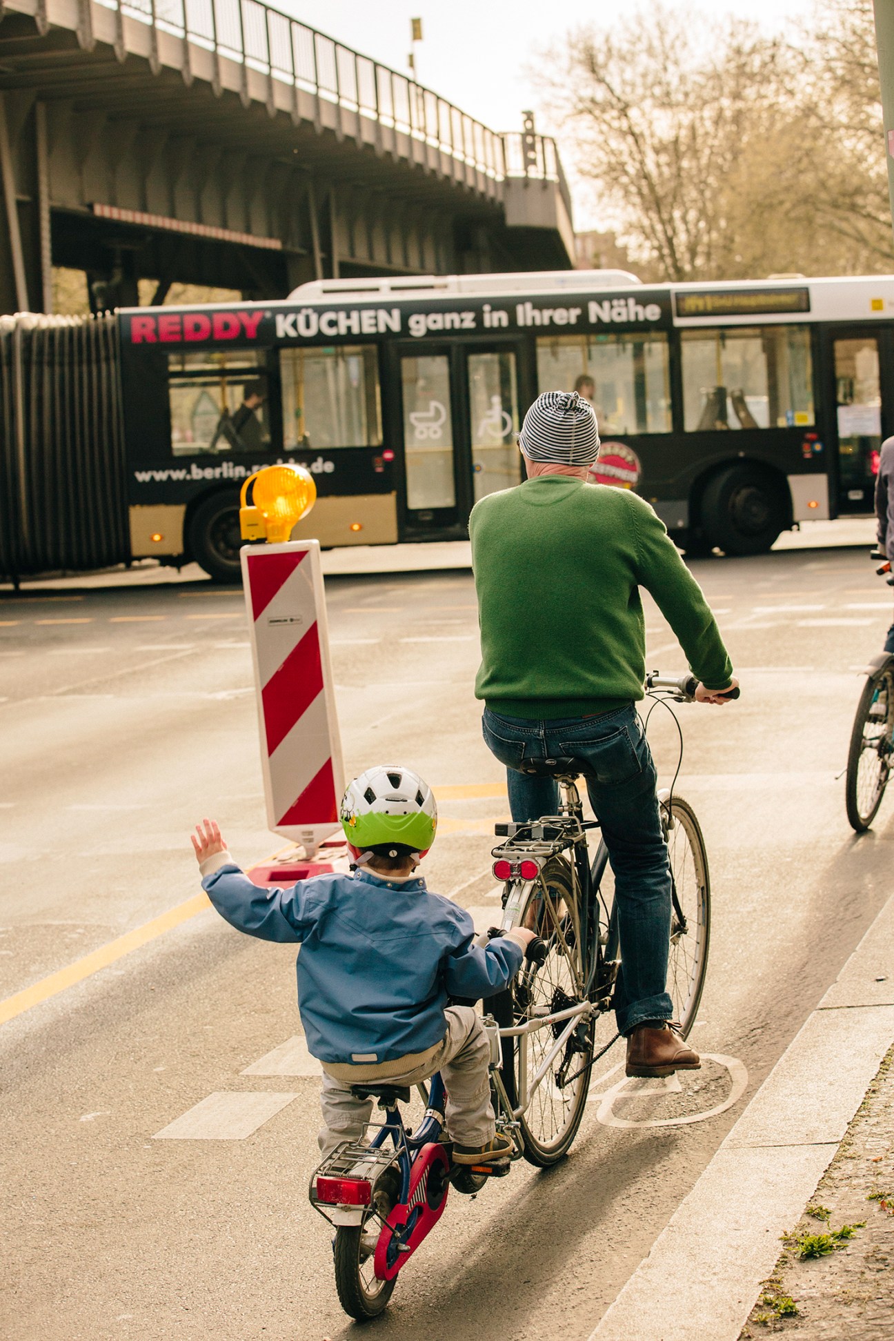 Adult and child cycling on a Berlin bike lane with a bus and bridge in the background during pandemic lockdown.