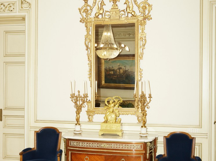 Ornate room with cream walls, gold decorative molding, antique wooden sideboard, blue velvet chairs, and gilded mirror