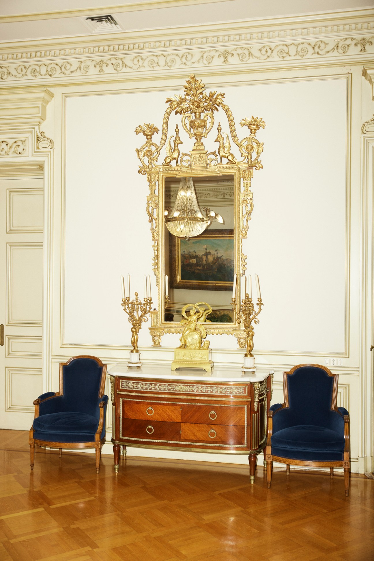 Ornate room with cream walls, gold decorative molding, antique wooden sideboard, blue velvet chairs, and gilded mirror