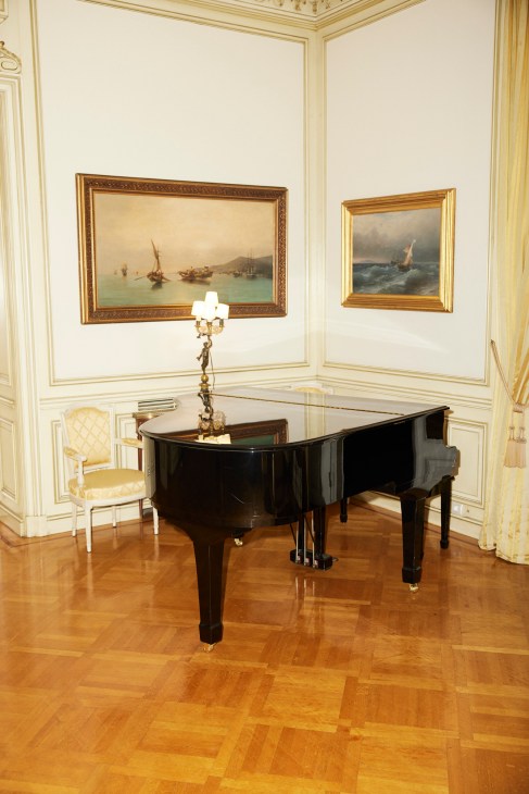 Black grand piano in ornate white room with gold trim, maritime paintings, and parquet flooring