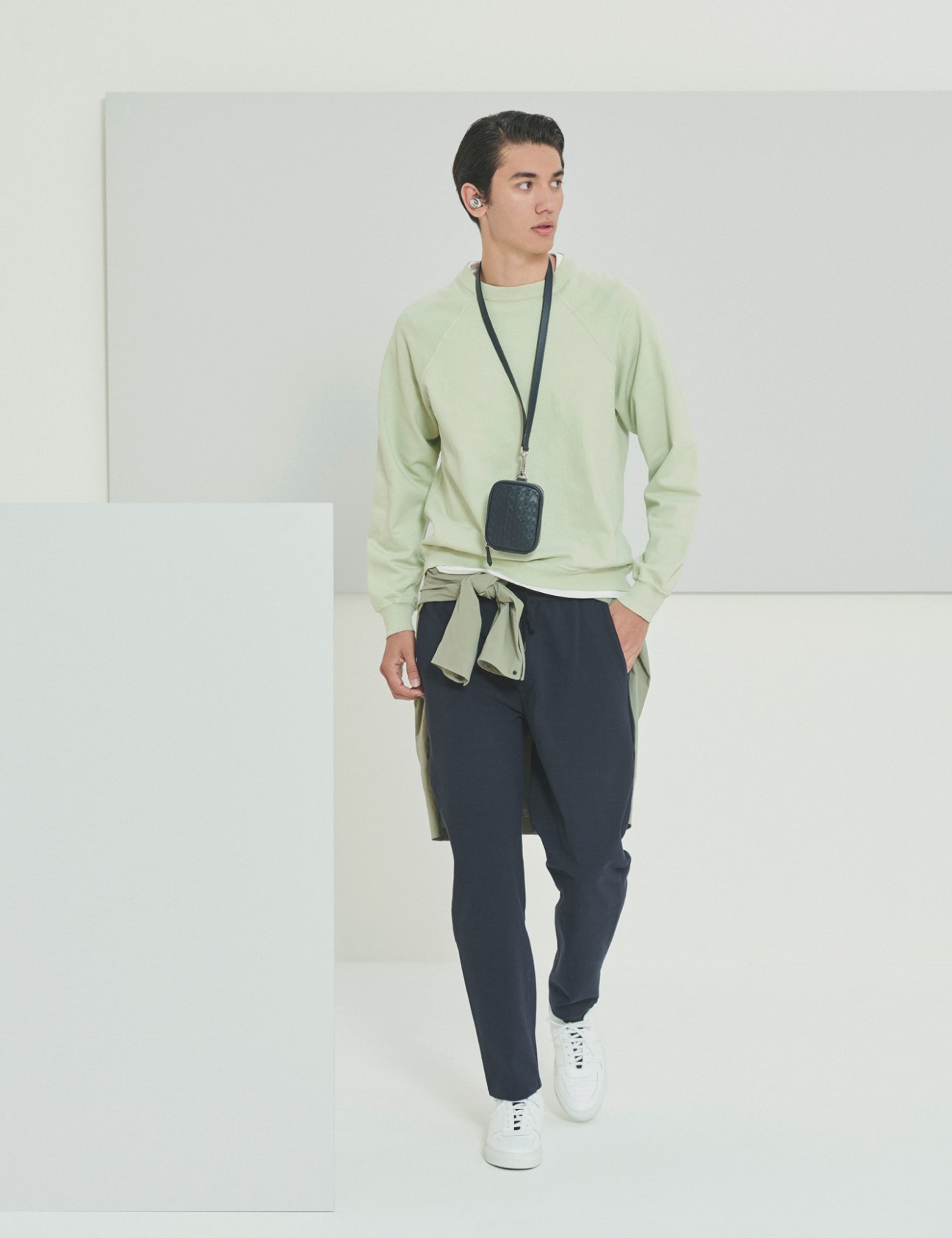 Model wearing light green sweatshirt, dark trousers, white sneakers and small crossbody bag