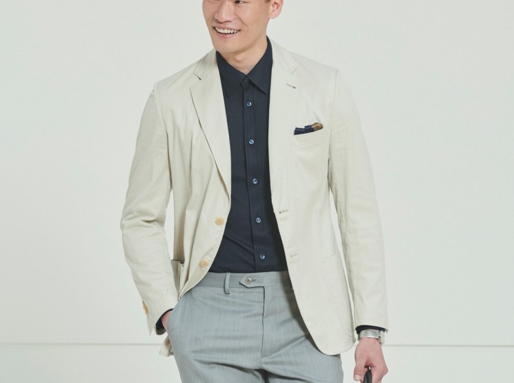 Model wearing cream blazer, navy shirt and grey trousers holding woven bag in studio