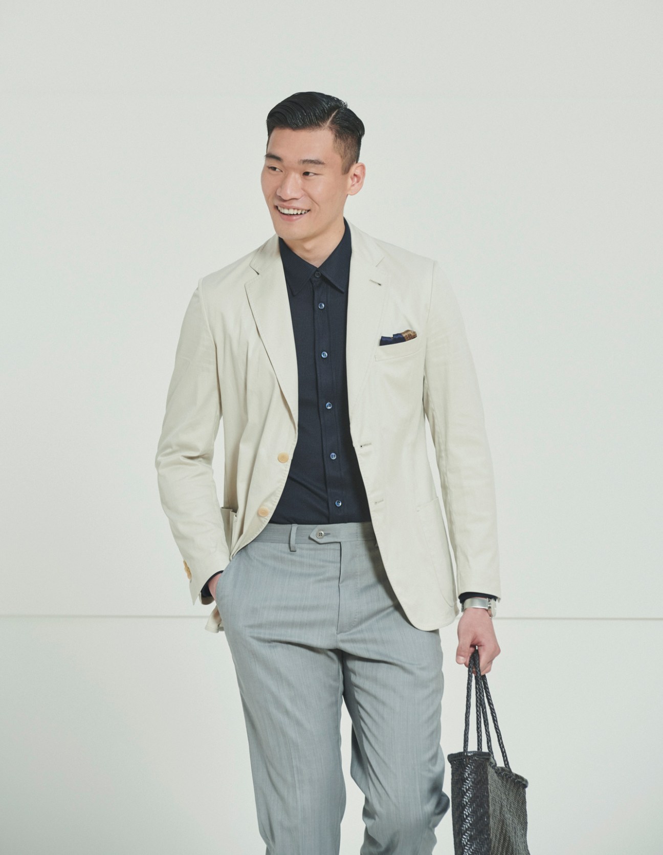 Model wearing cream blazer, navy shirt and grey trousers holding woven bag in studio