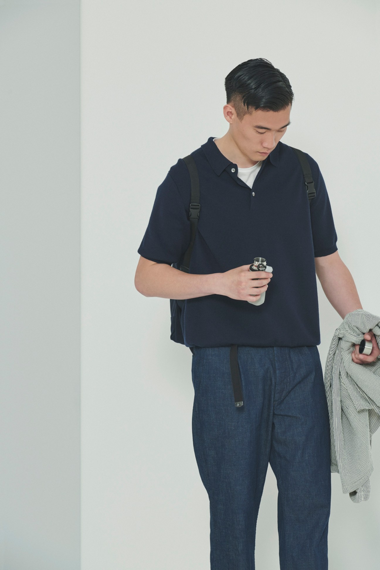 Model in navy polo shirt and blue trousers holding striped shirt in minimalist setting