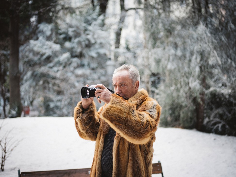 Elderly man in fur coat taking photograph with camera in snowy winter forest setting