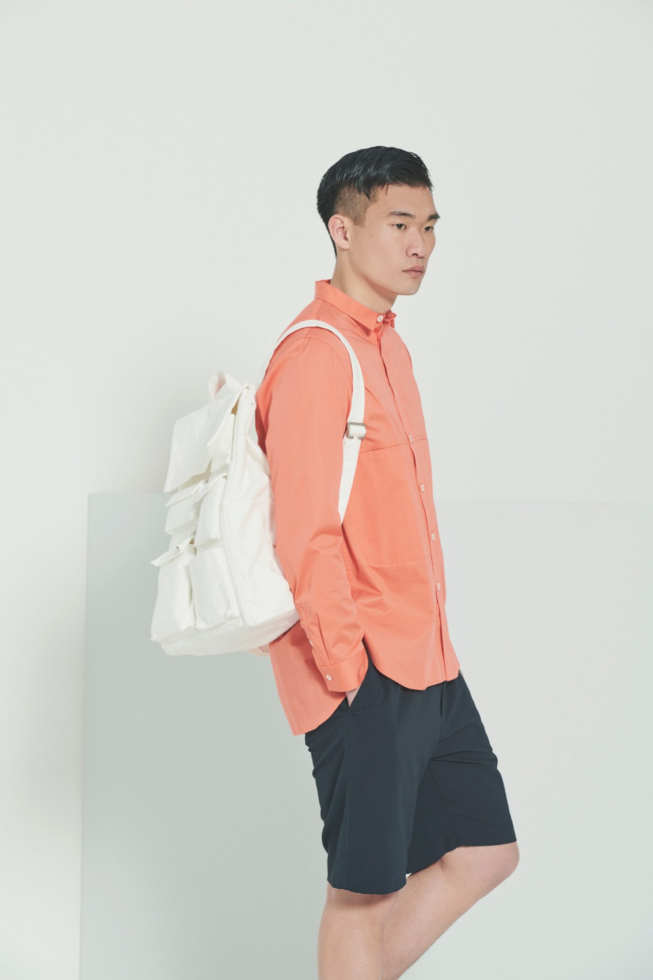 Model in coral shirt and navy shorts carrying white backpack against white background