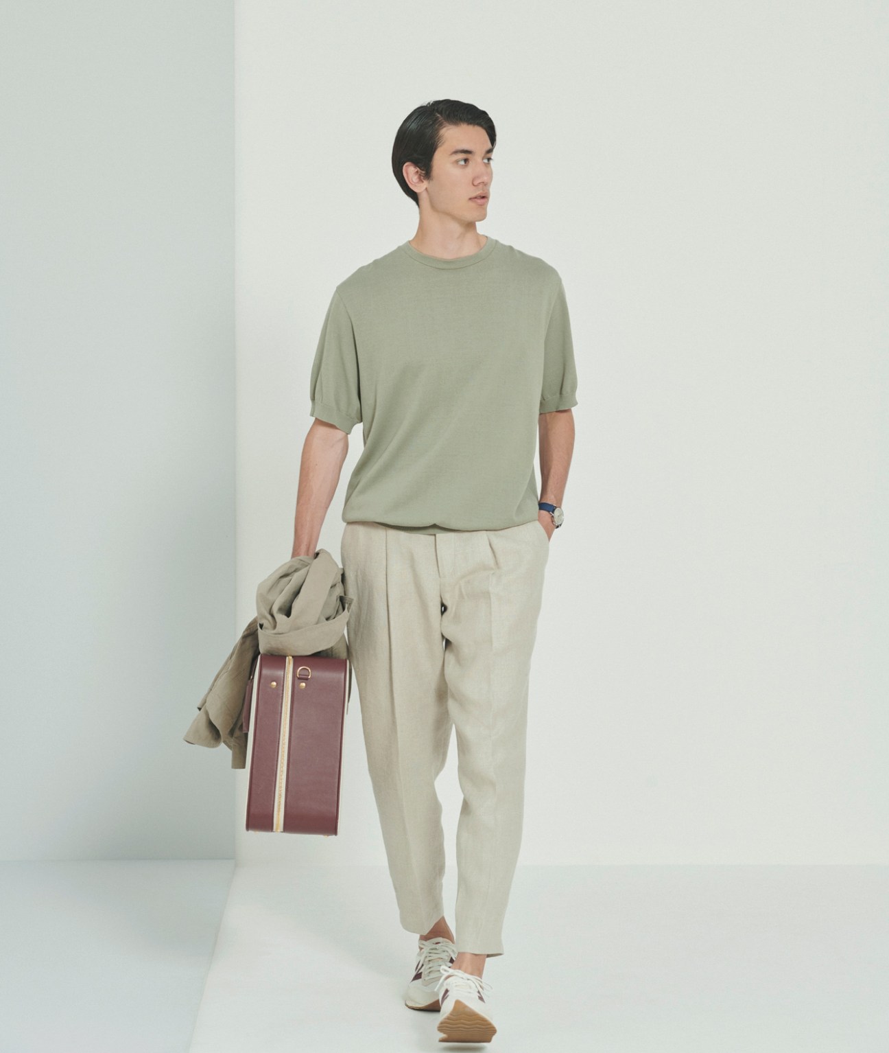 Model wearing sage green t-shirt and beige trousers, carrying brown leather bag and olive jacket