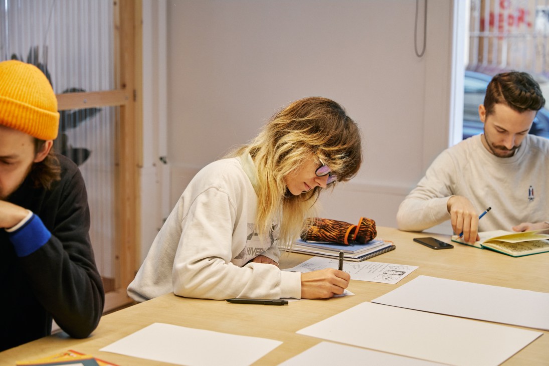Three students drawing and writing at wooden tables in a bright classroom setting, focused on their creative work