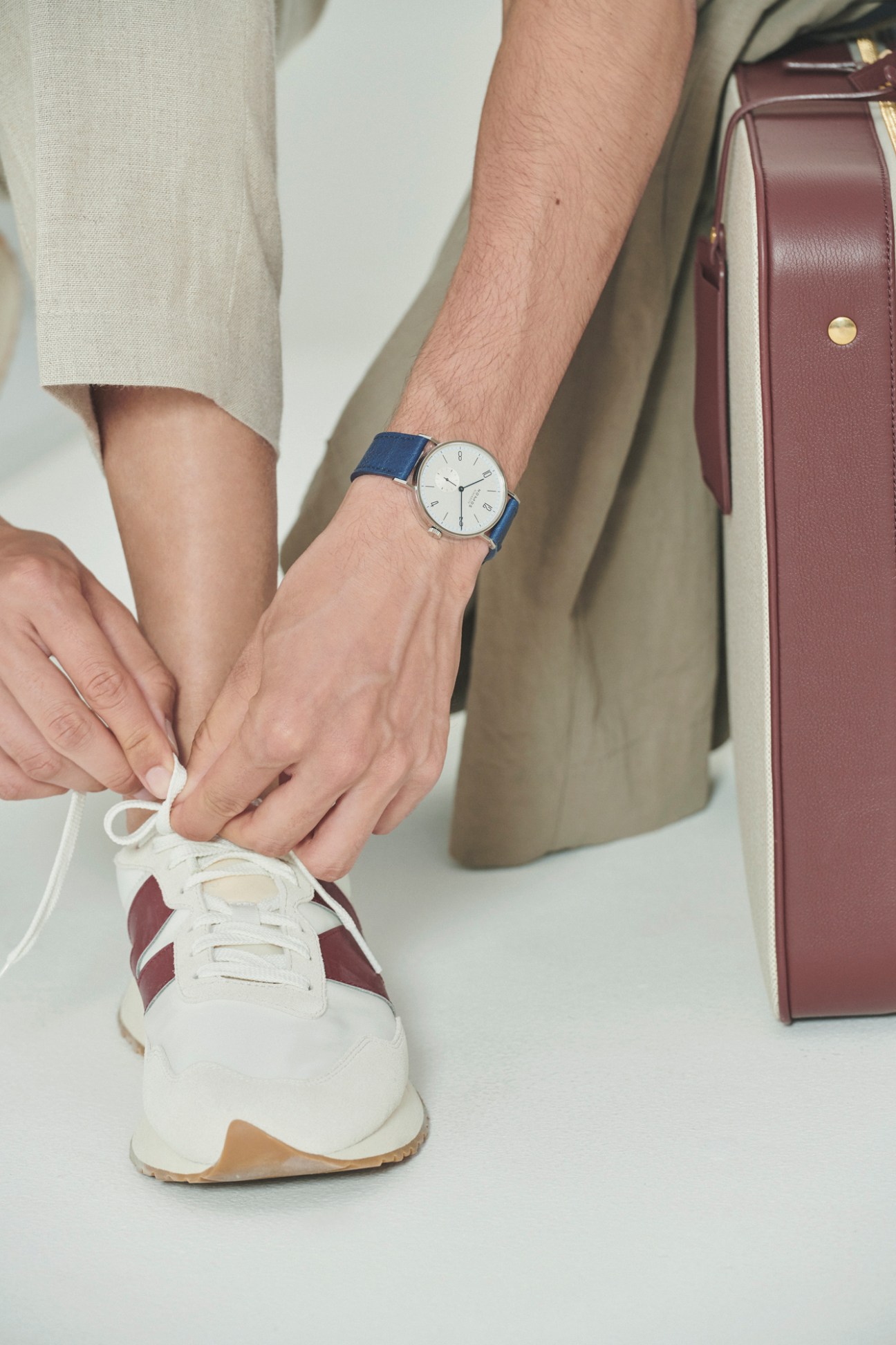 Person tying white New Balance trainers while wearing blue watch and burgundy bag