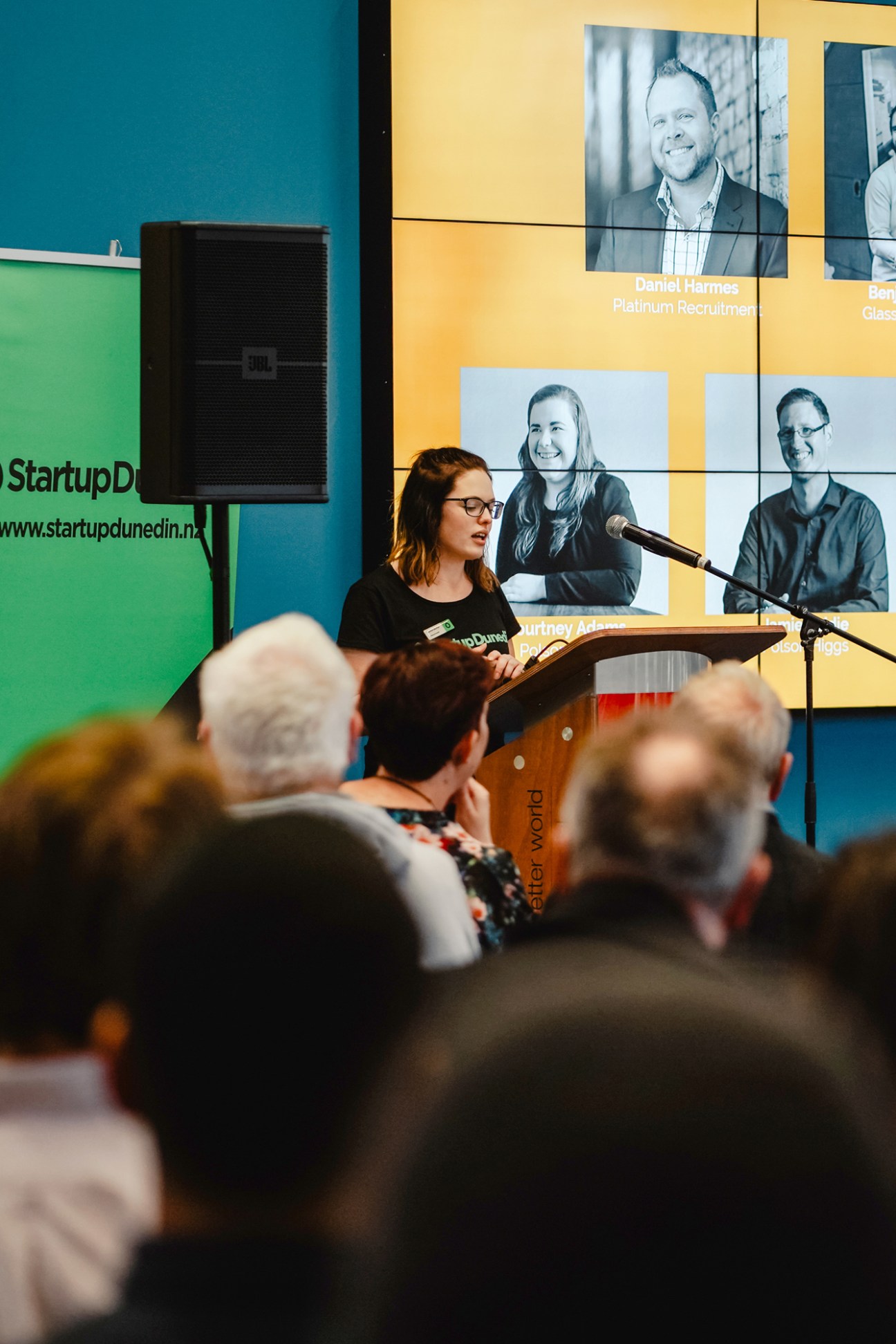 Woman speaking at podium in front of audience with portrait display behind her.