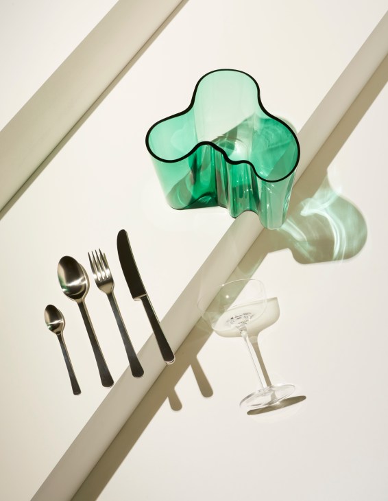 Green glass vase, black cutlery set, and clear champagne coupe arranged on white surface with dramatic shadows