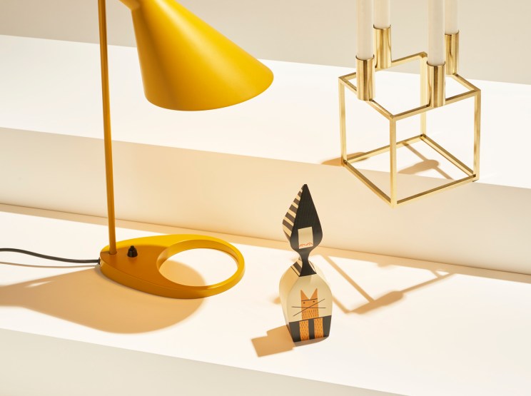 Yellow desk lamp, brass candle holder with lit candles, and wooden decorative figure on white stepped surfaces