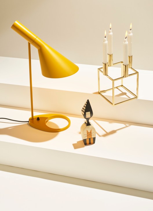 Yellow desk lamp, brass candle holder with lit candles, and wooden decorative figure on white stepped surfaces